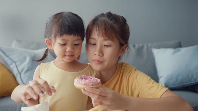 快乐快乐的亚洲家庭妈妈和蹒跚学步的小女孩在家里客厅的沙发上吃着甜甜圈，享受着快乐的放松。在一起的时间