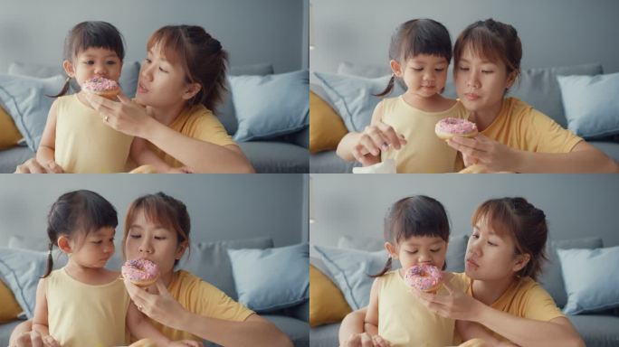快乐快乐的亚洲家庭妈妈和蹒跚学步的小女孩在家里客厅的沙发上吃着甜甜圈，享受着快乐的放松。在一起的时间