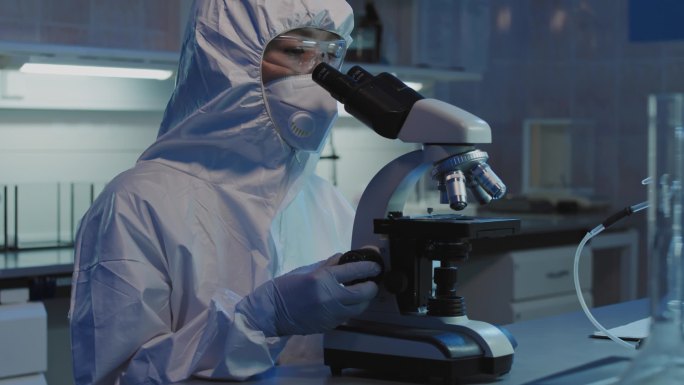 身着防护服、戴口罩、戴护目镜、戴着手套的女科学家在实验室用显微镜对着镜头看的慢镜头肖像画