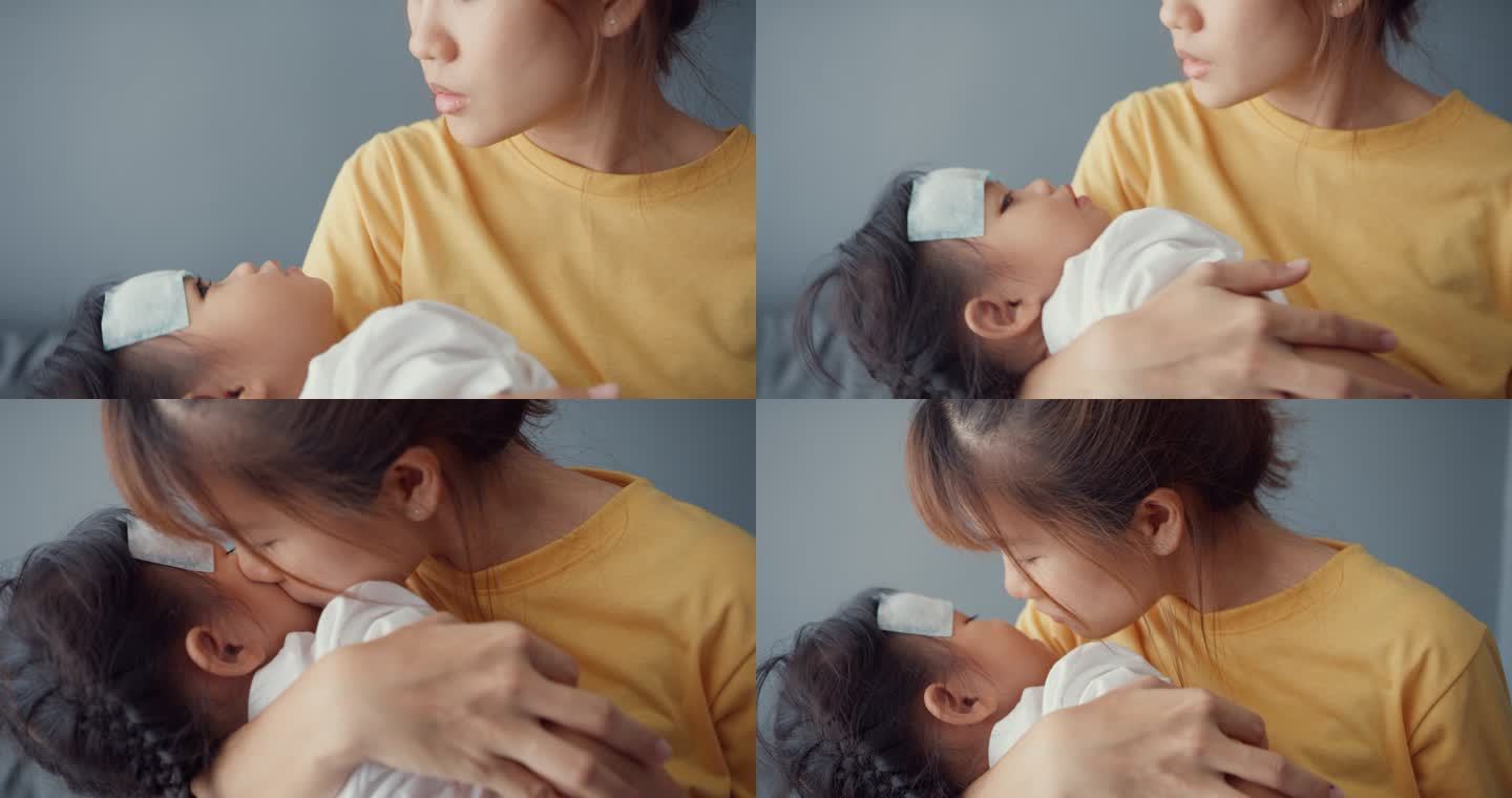 快乐快乐的亚洲家庭妈妈抱着生病的蹒跚学步的小女孩，在客厅里贴上解热凝胶，亲吻婴儿的额头。医疗保险、治