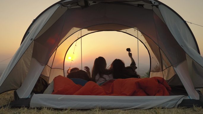 女孩朋友团体亚洲人在露营时躺在帐篷里醒来观看太阳升起，并从他们的动作摄像头拍摄视频，在社交媒体上向他