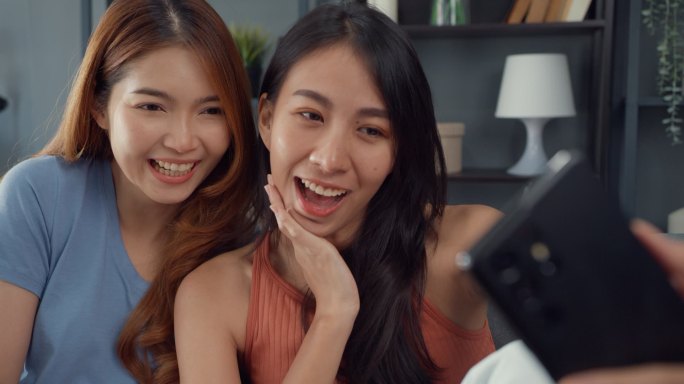 年轻的亚洲女人开心地笑着放松，在家里客厅里使用智能手机视频通话。快乐的室友女士与朋友和家人的视频会议