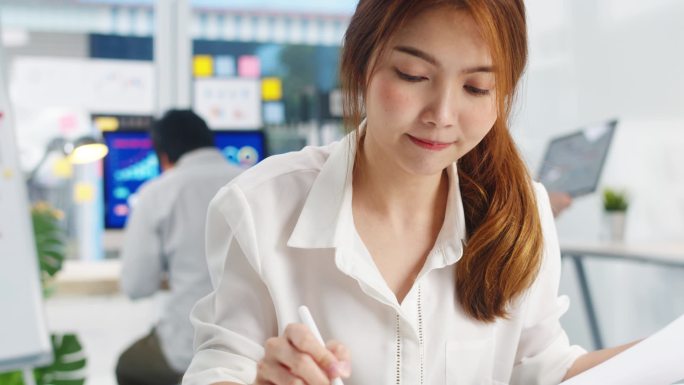 成功的亚洲执行董事年轻女商人智能休闲装的绘制、书写和使用带有数字平板电脑思维的钢笔在现代总公司的工作