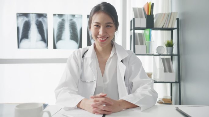 年轻、自信的亚洲女医生，身穿白色医疗制服，带着听诊器，看着相机，微笑着与医院里的病人进行视频会议。咨