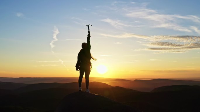 一位年轻的女登山者在日落时站在山顶上胜利地举起双手.