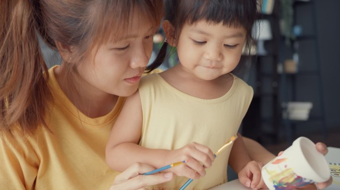 快乐快乐的亚洲家庭妈妈教蹒跚学步的小女孩画瓷壶在客厅的桌上玩乐放松。在一起度过的时间、社会距离、检疫