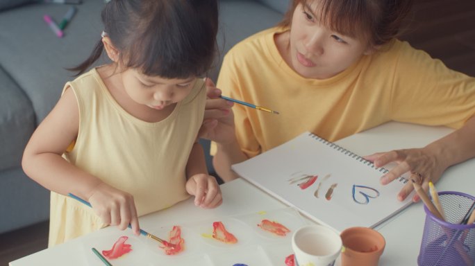 快乐快乐的亚洲家庭妈妈教蹒跚学步的小女孩画瓷壶在客厅的桌上玩乐放松。在一起度过的时间、社会距离、检疫