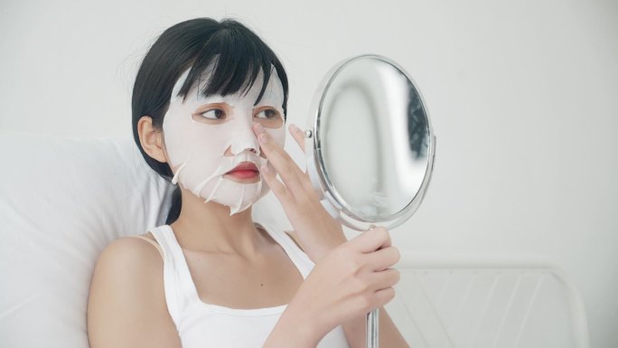 漂亮的年轻亚洲女人，戴着薄片面膜，在家里照镜子，漂亮的女孩用化妆品和化妆品，用皱纹护肤、护肤和保湿霜