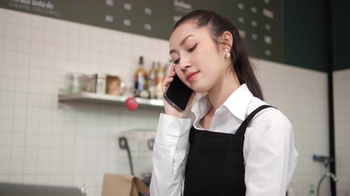 在咖啡店工作的亚洲妇女咖啡店员工。微笑的女服务员收银员在电话上向顾客订购咖啡和烘焙面包。小企业主和非