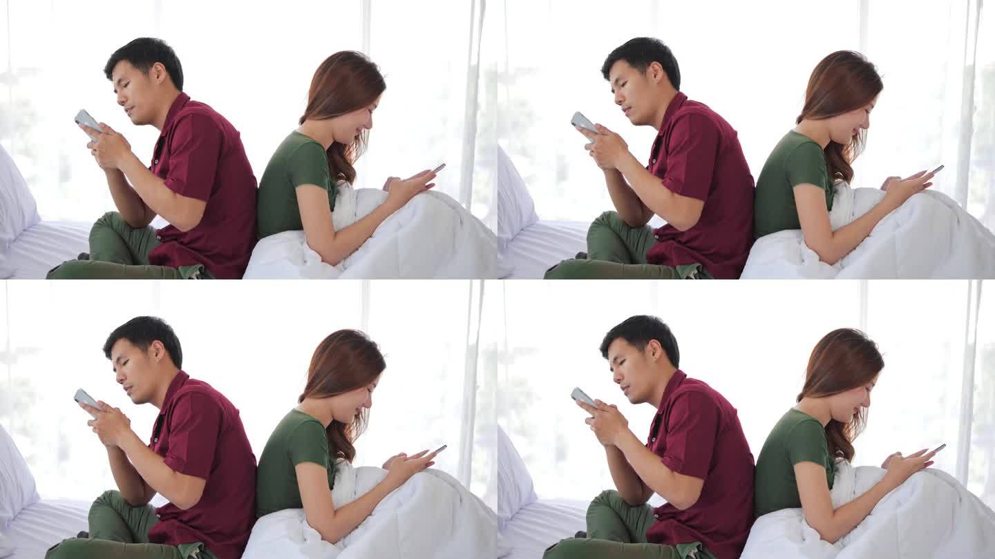 亚洲夫妇坐在床上，用手机发短信，用智能手机相互交流。一点也不不同，也不互相关心.
