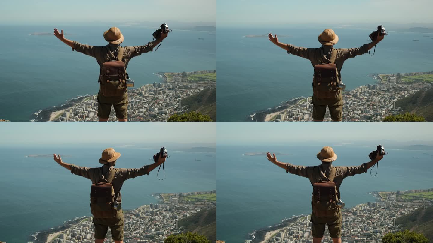 站在一座高山顶上的登山者凝视着并欣赏着全景落日。男人背着背包站在靠近大海的悬崖边上。男人举起双手在山