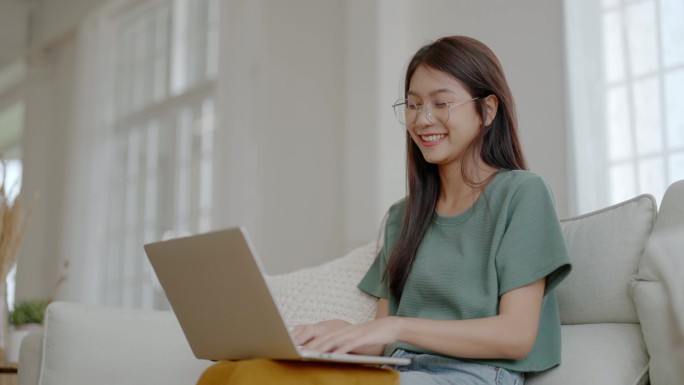 笑年轻的亚洲女人在家里的笔记本电脑上工作。通过计算机远程学习、在线教育和视频通话吸引亚洲女性