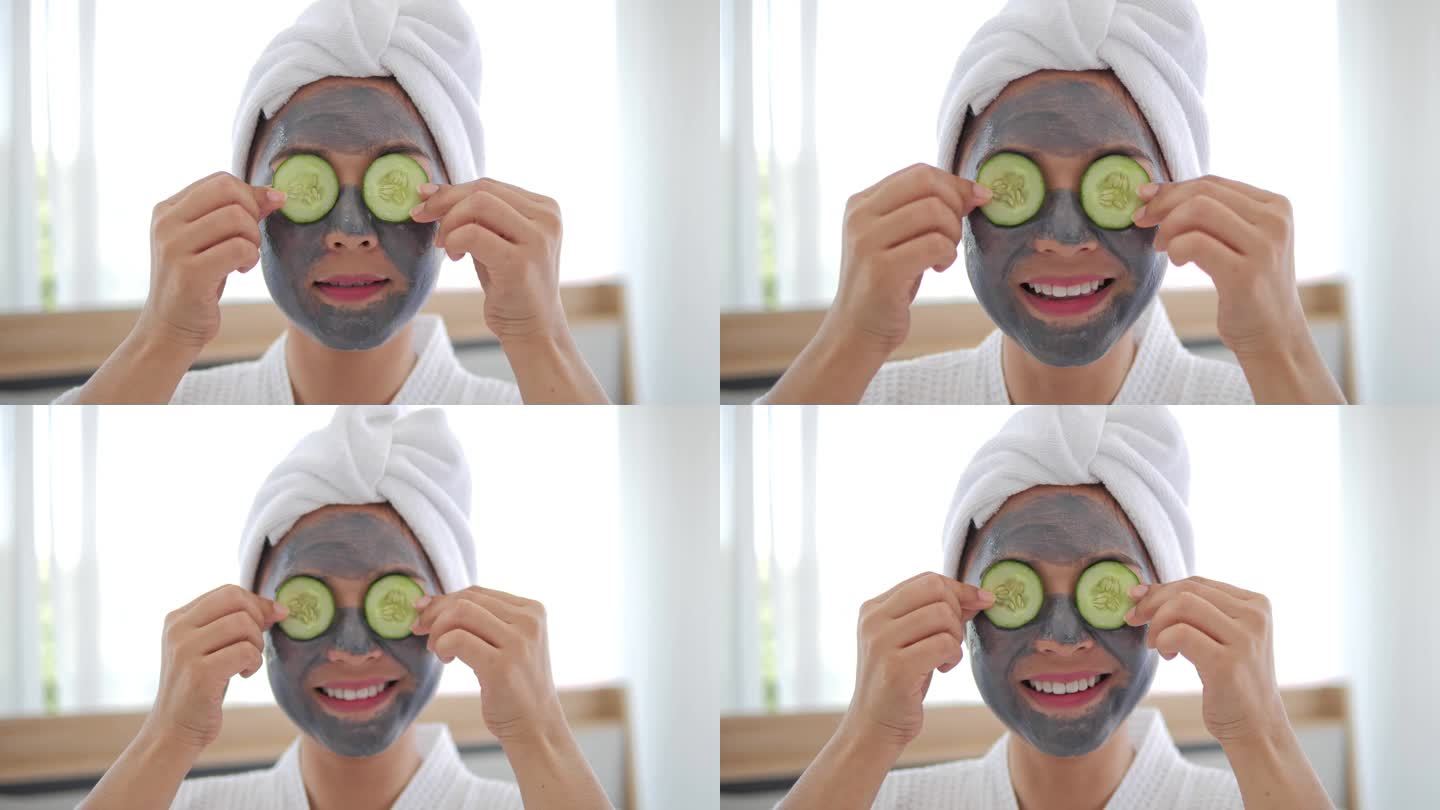 脸上戴着灰色面具的亚洲女人正在把几片黄瓜放在眼睛上，微笑着看着相机。水疗美容程序在家里为女孩。护肤、