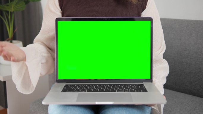 女人在家里的沙发上展示带有绿色屏幕和彩色键的笔记本电脑。显示绿色屏幕并与相机对话