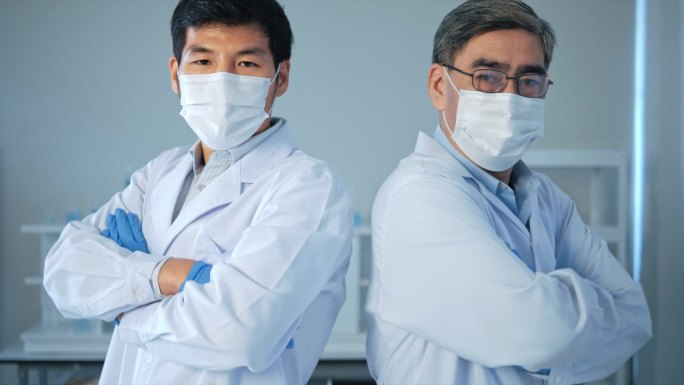 两名戴口罩的亚洲男性医生交叉着双臂，在实验室试管中看着摄像机检测蓝色液体。医疗科学保健概念