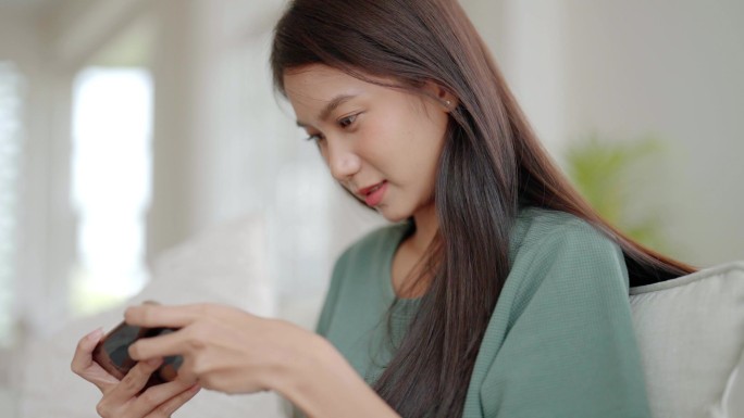 快乐的年轻亚洲女人在家里舒服的沙发上放松，在智能手机上玩网络游戏，微笑的女孩用手机聊天，在电子产品上