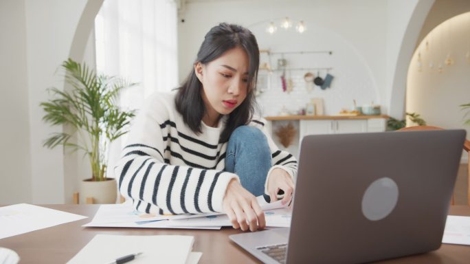 年轻的亚洲女性自由撰稿人专注于笔记本电脑的演示工作，以及在家里厨房与公司进行远程会议的财务项目。女生