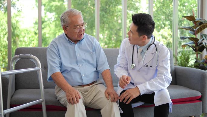 亚洲男医生身穿白衣检查膝盖的老年男子坐在沙发上，老年人保健支持理念.