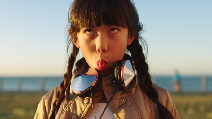 亚洲儿童，脸或面部表情和舌头通过海滩，海洋或大海和凉爽，时尚或时尚的音乐耳机。巴厘岛自然环境中的人物