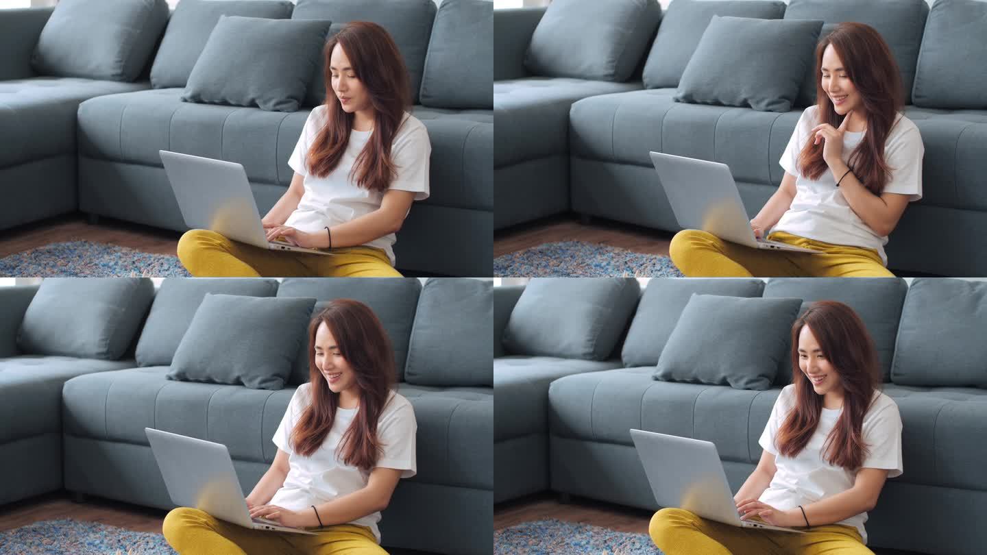 微笑的年轻亚洲妇女在家沙发下面工作。坐在笔记本电脑上的亚洲妇女看着屏幕打字信息。疏远在家在线工作的概