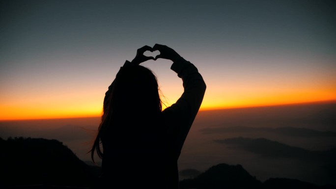 日出时手放在山顶上慢吞吞地做着心脏的女人的轮廓。快乐的年轻女人轮廓手握心形在地平线上.