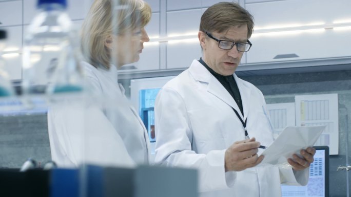 男性和女性首席科学家步行穿过现代实验室，讨论未来的可能性。计算机在她的办公桌上年轻女助理工程。