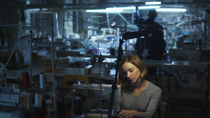 年轻女子制衣工人是在夜班期间在一家服装厂缝纫而灯都关掉在背景中.