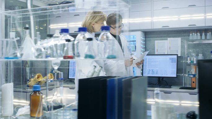 女性和男性的高级科学家步行穿过繁忙的现代实验室。他们有关于文档的讨论。