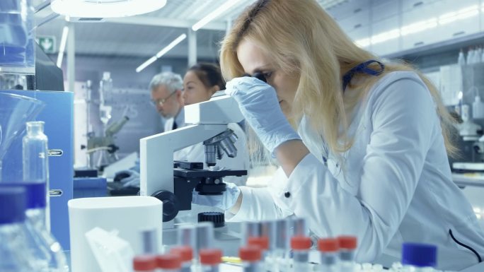 女性研究的科学家在生物样品在显微镜下看起来。她和她的同事在一个大的现代实验室工作.