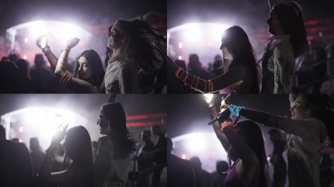 两个女孩在露天派对上用闪烁的灯光和激光跳舞。朋友们一个接一个地站着。失焦人群和dj舞台的背景。