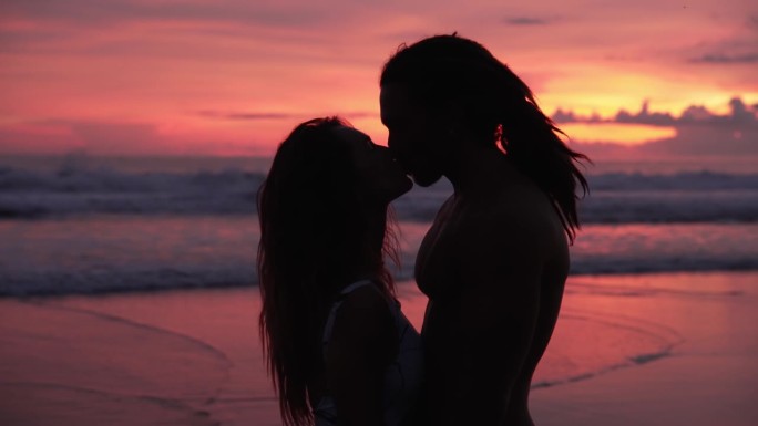 一对相爱的人在海边，日落和海水。现代高加索家庭的两个恋人放松地看着对方，亲吻。漂亮女人和有吸引力的英