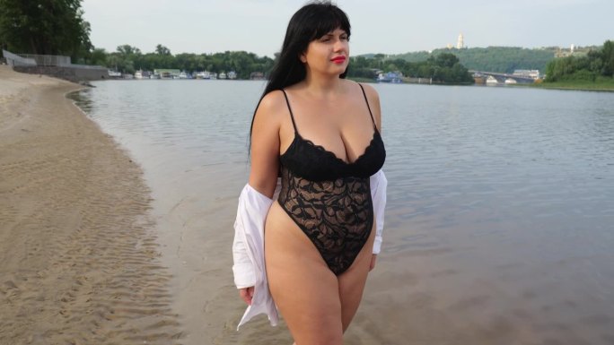 一个30岁的大码模特穿着黑色内衣和白色衬衫走在海边。暑假在海滩上，胖的黑发女人，长头发