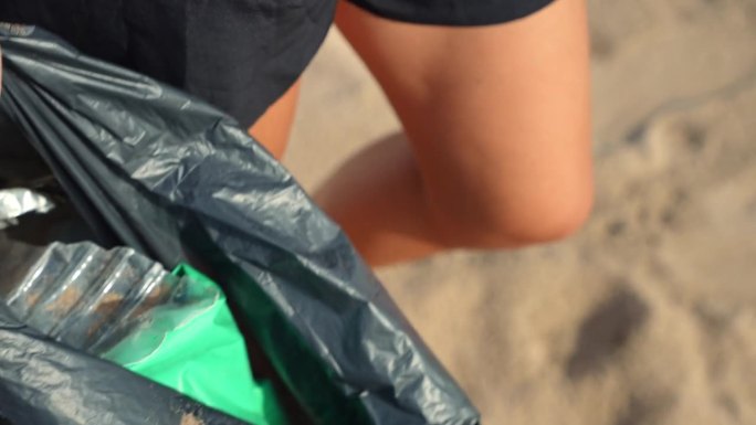 年轻女子戴着防护手套从海滩上捡垃圾，把垃圾放进垃圾袋里