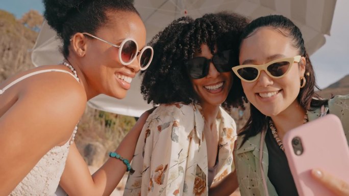 女性朋友，在度假或野餐时，戴着太阳镜，撅着嘴，在沙滩上自拍。多元化女子组合在夏天把智能手机放在伞下，