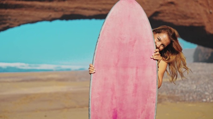 一名女子在阳光明媚的海滩上拿着粉红色的冲浪板摆姿势，脸上带着快乐和顽皮的表情