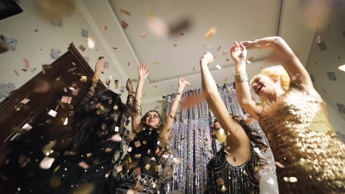 四个年轻迷人的女人在闪闪发光的礼服欢呼在飞舞的彩纸在生日聚会上跳舞。缓慢的运动。