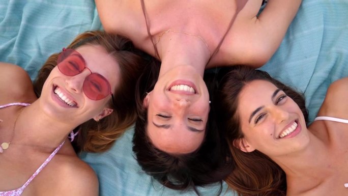 高角度拍摄的三个开朗的女性躺在沙滩浴巾上，看着镜头开心的样子。