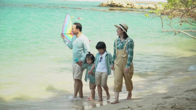 快乐的亚洲家庭散步和手牵风筝在海滩上一起旅行在假期暑假。妈妈爸爸女儿儿子暑假旅行。穿着相似的服装。