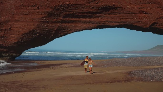 年轻的冲浪夫妇在沙滩上浪漫地漫步，海浪拍打着沙滩，背景是天然的红砂岩拱门