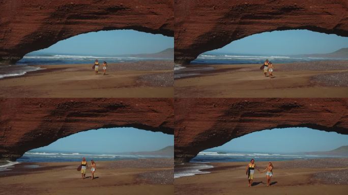年轻的冲浪夫妇在沙滩上浪漫地漫步，海浪拍打着沙滩，背景是天然的红砂岩拱门