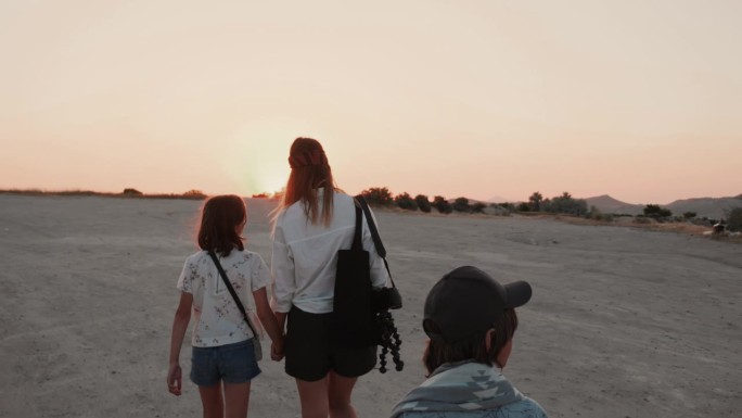镜头跟随年轻快乐的母亲，十几岁的儿子和女儿在日落时分沿着海滩散步，一起享受暑假。