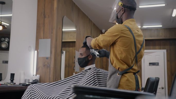 在covid-19大流行期间，理发师在理发店戴着医用口罩和屏幕剃须的非洲裔美国人