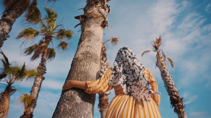 穆斯林妇女戴着头巾，触摸着棕榈树，在蓝天下行走