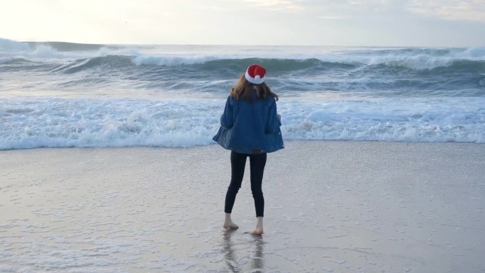一个戴着红色圣诞老人帽的女孩或年轻女子在大西洋的海滩上玩耍，奔跑和转身。圣诞节或新年假期的概念。4K