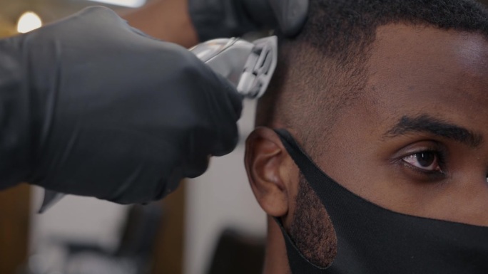 戴着医用口罩的男子在理发店理发时，手戴手套使用剃须机的特写