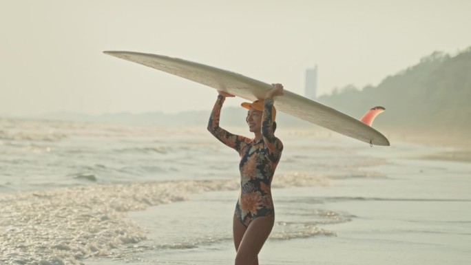日落时分，女冲浪者带着冲浪板在海滩上散步，慢动作