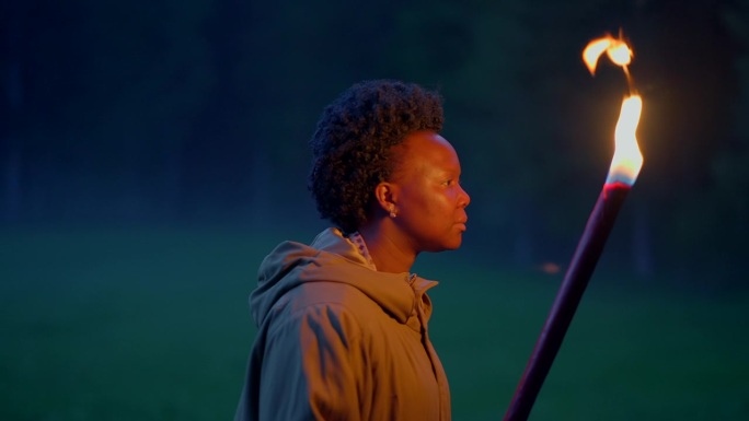 黑人女性在自然森林景观中行走，用燃烧的火炬光发现户外的黑暗