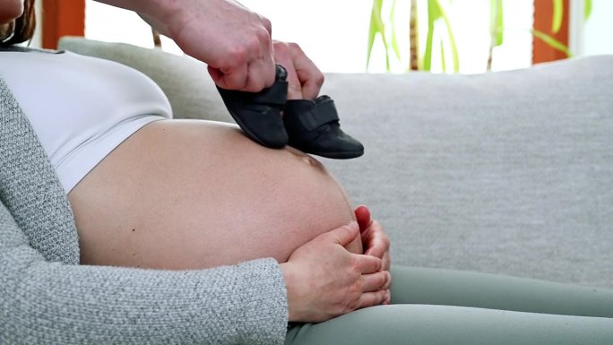 在家里的公寓里，坐在沙发上的孕妇的圆肚子上，爸爸穿着婴儿鞋做了一个“太空步”。妊娠晚期-第39周。侧