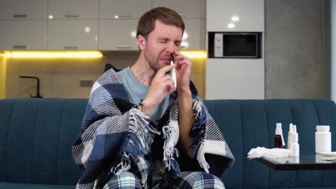 一名男子在家里的沙发上用鼻喷雾剂治疗流鼻涕和鼻塞。疾病的治疗。鼻炎、鼻窦炎、感冒。对滴剂的依赖