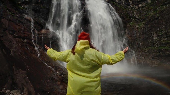 穿着黄色雨衣的年轻女孩徒步旅行者走到高地的瀑布前，举起双手，享受自然和生活。在山中旅行，在旅途中冒险